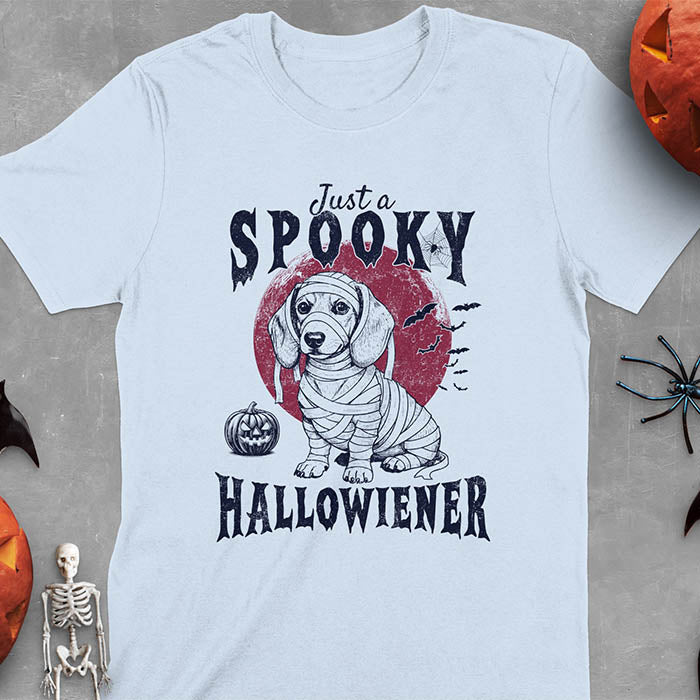 Light blue t-shirt featuring a cute dachshund puppy wrapped in mummy bandages with text 'Just a Spooky Hallowiener', jack-o'-lantern pumpkin, red moon and flying bats in background