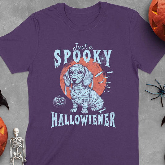Purple t-shirt featuring a cute dachshund puppy wrapped in mummy bandages with text 'Just a Spooky Hallowiener', jack-o'-lantern pumpkin, red moon and flying bats in background