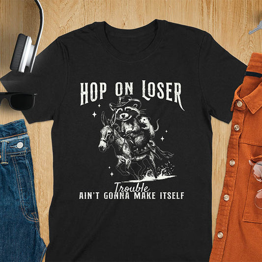 A black tshirt featuring a raccoon riding a donkey with an opossum clinging to its back. Text reads Hop On Loser and Trouble Ain't Gonna Make Itself.