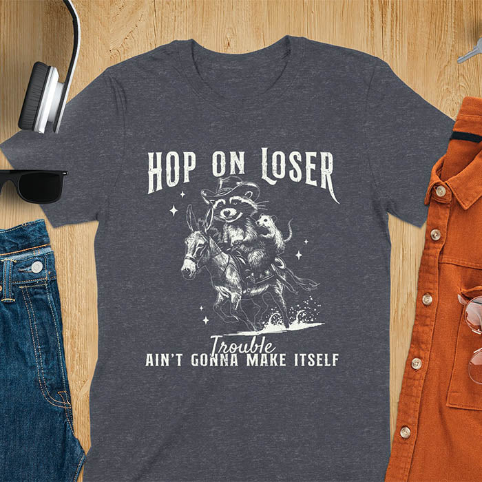 A dark heather tshirt featuring a  raccoon riding a donkey with an opossum clinging to its back. Text reads Hop On Loser and Trouble Ain't Gonna Make Itself.