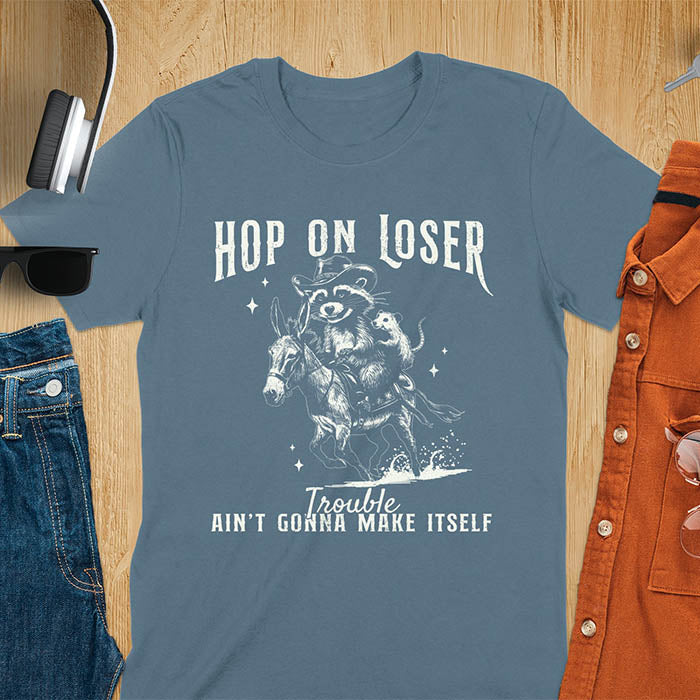 A indigo blue tshirt featuring a raccoon riding a donkey with an opossum clinging to its back. Text reads Hop On Loser and Trouble Ain't Gonna Make Itself.