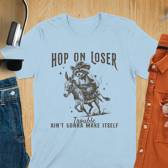 A light blue tshirt featuring a raccoon riding a donkey with an opossum clinging to its back. Text reads Hop On Loser and Trouble Ain't Gonna Make Itself.