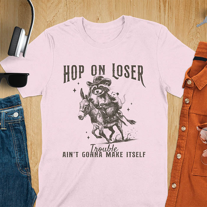 A light pink tshirt featuring a  raccoon riding a donkey with an opossum clinging to its back. Text reads Hop On Loser and Trouble Ain't Gonna Make Itself.