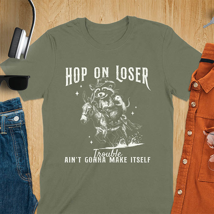A military green tshirt featuring a raccoon riding a donkey with an opossum clinging to its back. Text reads Hop On Loser and Trouble Ain't Gonna Make Itself.