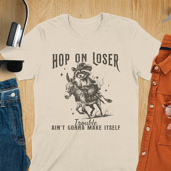 A sand color tshirt featuring a raccoon riding a donkey with an opossum clinging to its back. Text reads Hop On Loser and Trouble Ain't Gonna Make Itself.