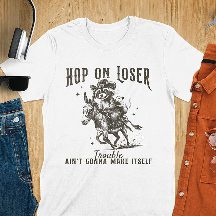 A white tshirt featuring a raccoon riding a donkey with an opossum clinging to its back. Text reads Hop On Loser and Trouble Ain't Gonna Make Itself.