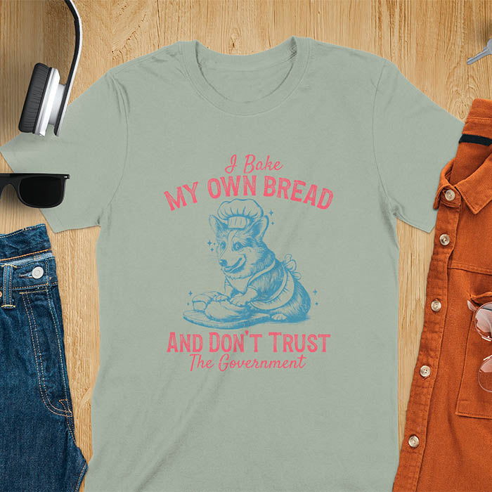 A t-shirt with a corgi wearing a chef's hat and apron, kneading dough. Text: "I Bake MY OWN BREAD" above and "AND DON'T TRUST The Government" below. This retro sarcastic tee is a funny meme shirt and funny dog shirt, a corgi shirt for a corgi lovers gift. An animal lovers tshirt and dog lover gift, a dog mom shirt or pet dad shirt. A dog person tee and pet dog owner gift, one of the funny bakers gifts, a gift for mom or her.