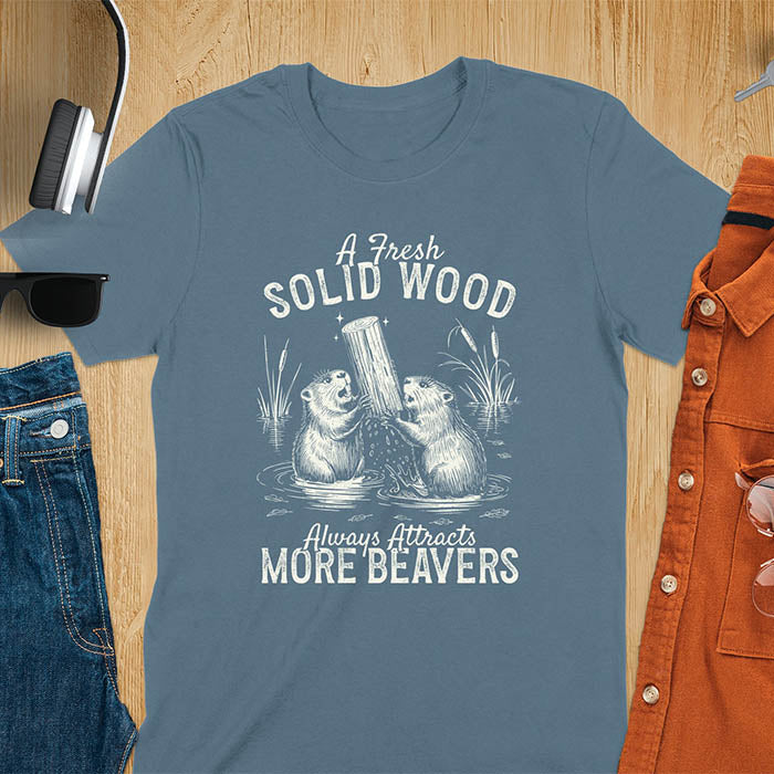 Indigo blue t-shirt with vintage-style illustration of two beavers in water fighting over a wooden log, featuring text 'A Fresh Solid Wood Always Attracts More Beavers' in rustic lettering 
