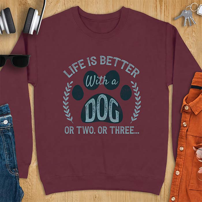 Maroon sweatshirt with a graphic of a large paw print framed by a laurel wreath on either side and the text Life Is Better With a Dog Or Two Or Three