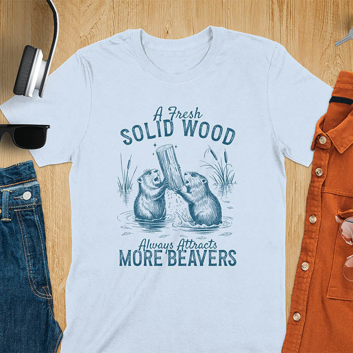 Light blue t-shirt with vintage-style illustration of two beavers in water fighting over a wooden log, featuring text 'A Fresh Solid Wood Always Attracts More Beavers' in rustic lettering 