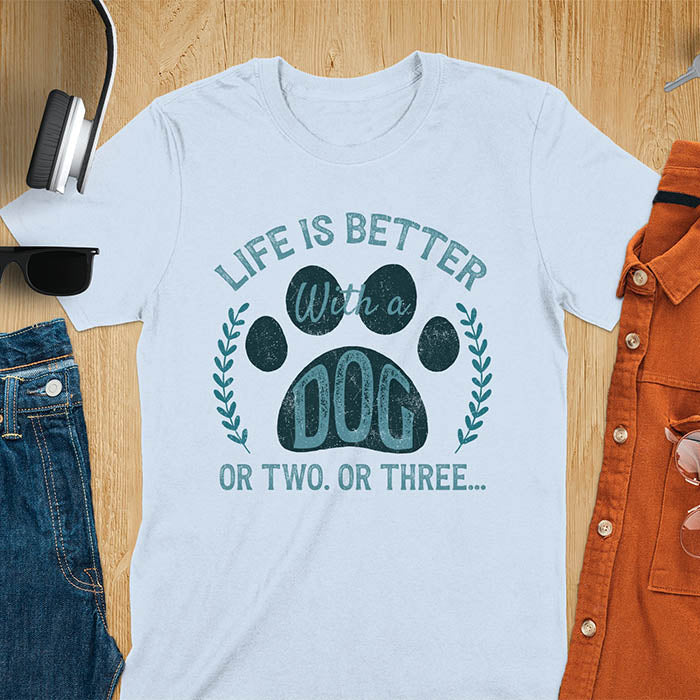Light blue t-shirt featuring large paw print with text inside surrounded by laurel wreath, text reads Life Is Better With A Dog Or Two Or Three