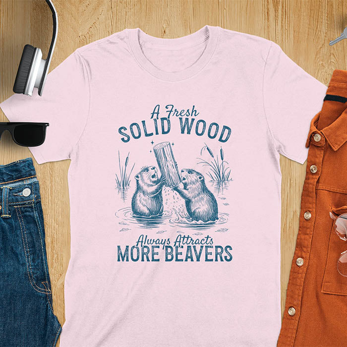 Light pink t-shirt with vintage-style illustration of two beavers in water fighting over a wooden log, featuring text 'A Fresh Solid Wood Always Attracts More Beavers' in rustic lettering 