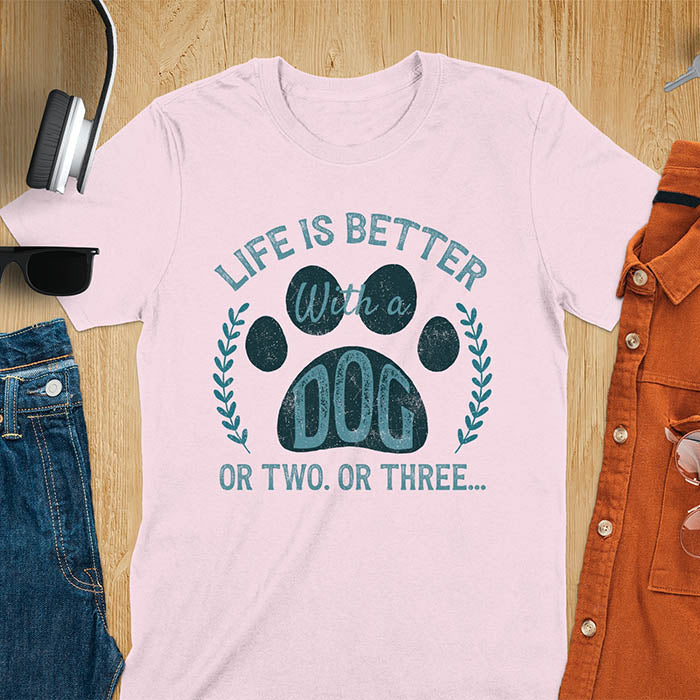 Light pink t-shirt featuring large paw print with text inside surrounded by laurel wreath, text reads Life Is Better With A Dog Or Two Or Three