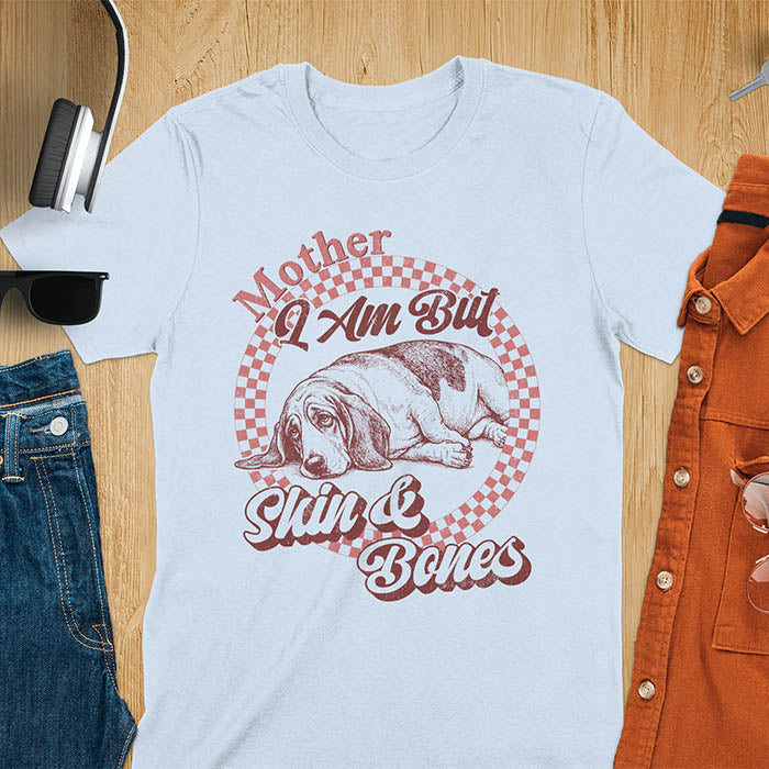 Light blue t-shirt with a vintage graphic of a basset hound lying down inside a retro checkered circle, with the text Mother I Am But Skin and Bones