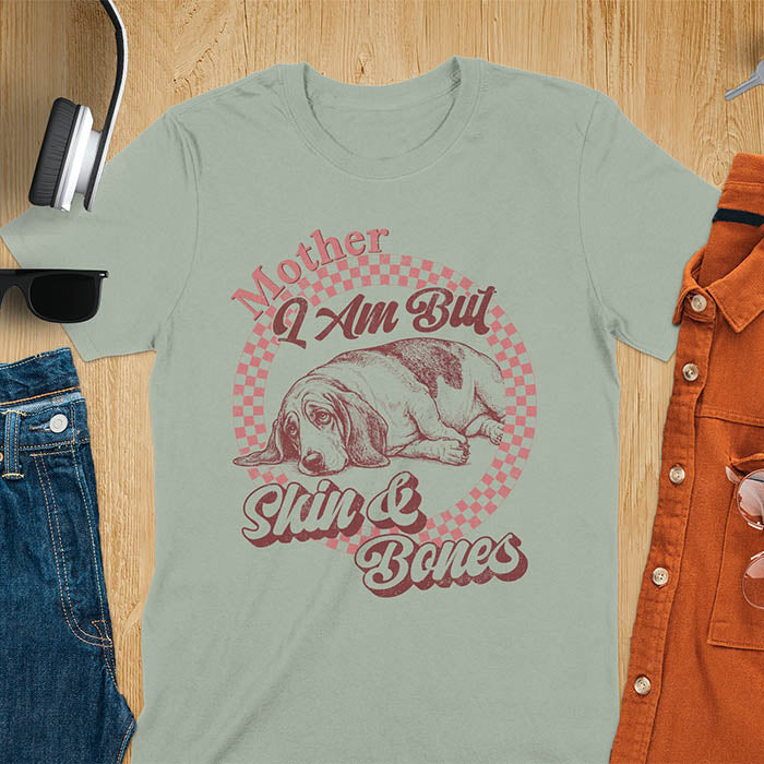Sage t-shirt with a vintage graphic of a basset hound lying down inside a retro checkered circle, with the text Mother I Am But Skin and Bones