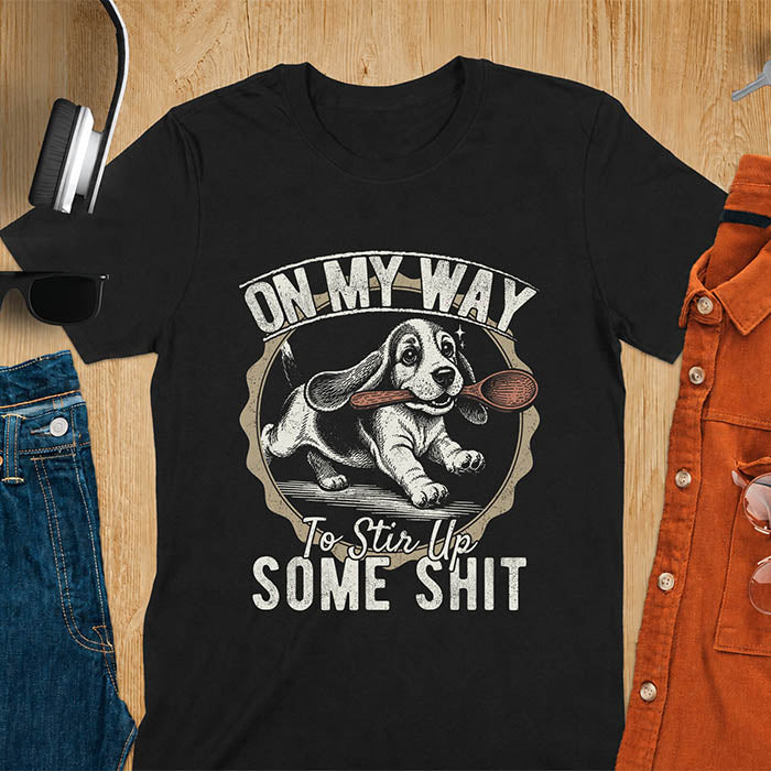 Black graphic tee with vintage stamp illustration of a basset hound running with a wooden spoon, text reads On My Way To Stir Up Some Shit