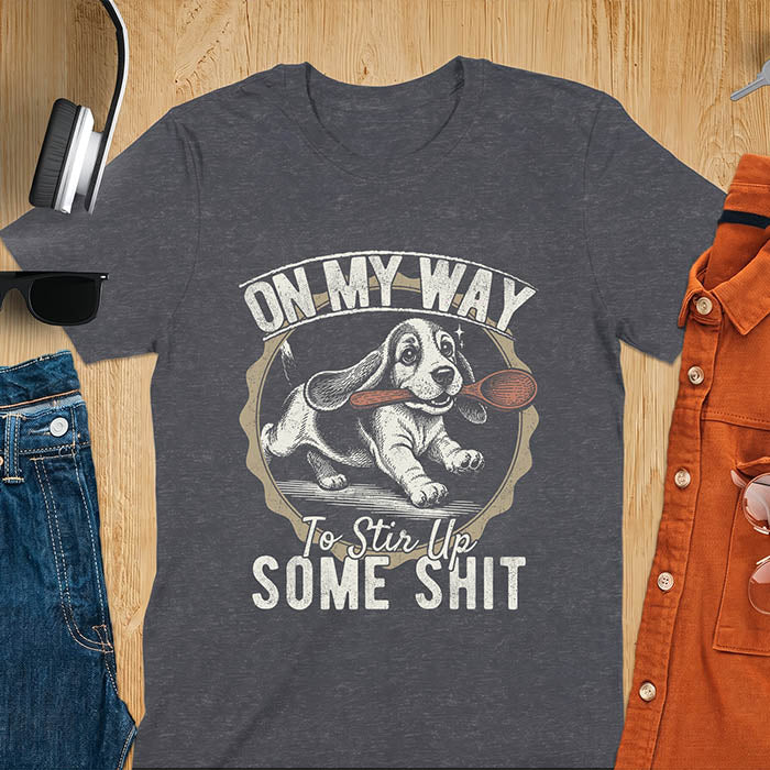 Dark heather graphic tee with vintage stamp illustration of a basset hound running with a wooden spoon, text reads On My Way To Stir Up Some Shit
