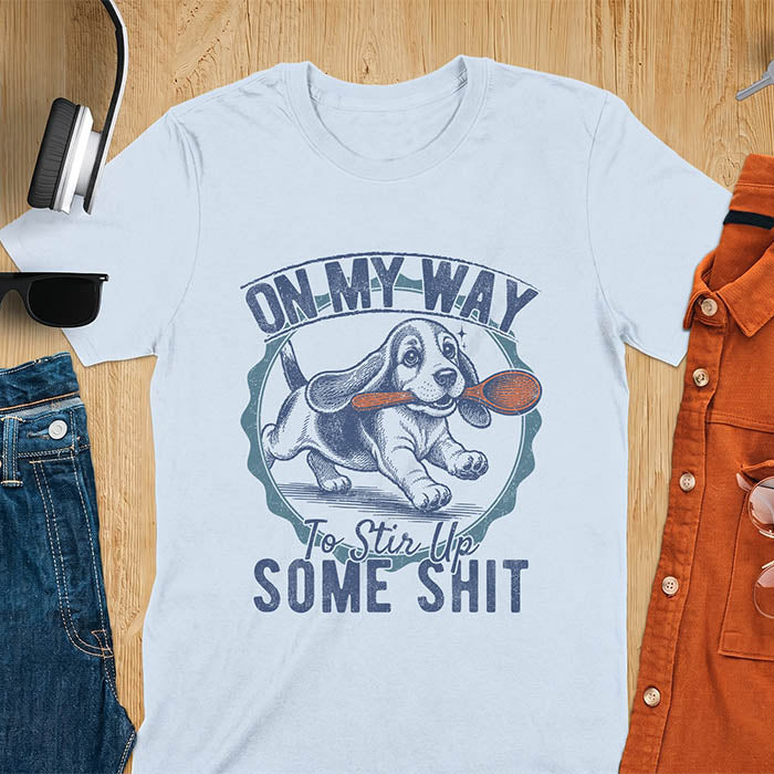 Light blue graphic tee with vintage stamp illustration of a basset hound running with a wooden spoon, text reads On My Way To Stir Up Some Shit