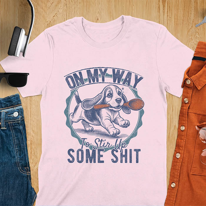 Light Pink graphic tee with vintage stamp illustration of a basset hound running with a wooden spoon, text reads On My Way To Stir Up Some Shit