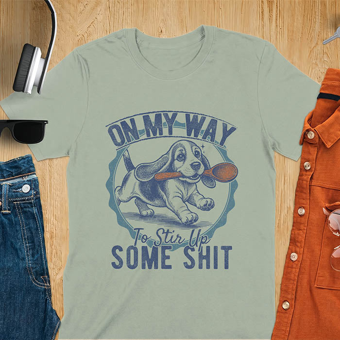 Sage green graphic tee with vintage stamp illustration of a basset hound running with a wooden spoon, text reads On My Way To Stir Up Some Shit