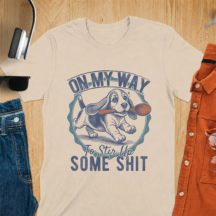 Sand color graphic tee with vintage stamp illustration of a basset hound running with a wooden spoon, text reads On My Way To Stir Up Some Shit