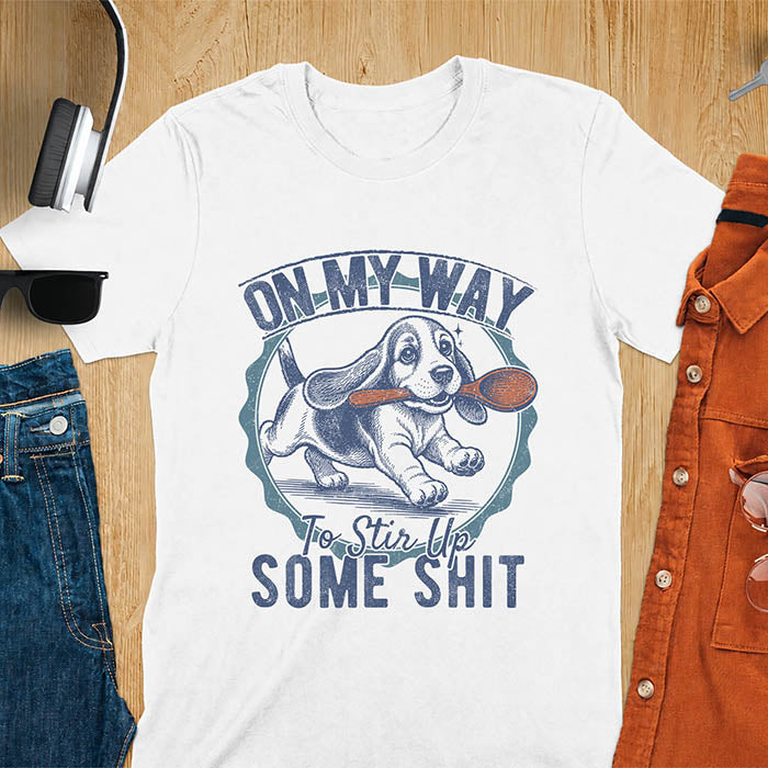 White graphic tee with vintage stamp illustration of a basset hound running with a wooden spoon, text reads On My Way To Stir Up Some Shit
