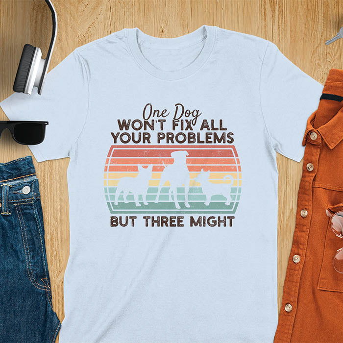 Light blue t-shirt with text 'One dog won't fix all your problems but three might' and three dog silhouettes on vintage sunset stripes