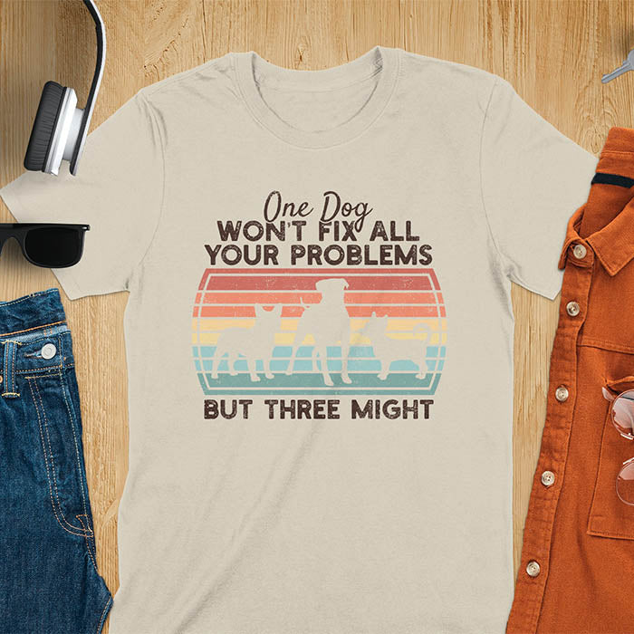 Sand color blue t-shirt with text 'One dog won't fix all your problems but three might' and three dog silhouettes on vintage sunset stripes