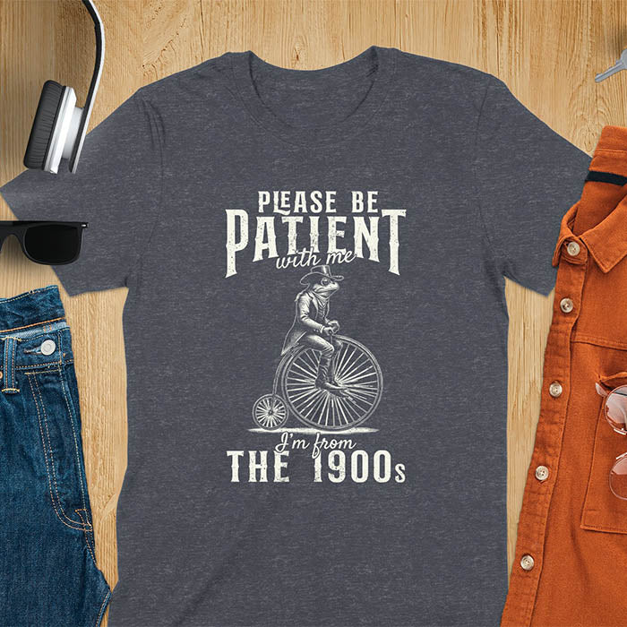 A dark heather t-shirt with a frog riding a penny-farthing bicycle. Text: "PLEASE BE PATIENT with me" above and "I'm from THE 1900s" below. This The 1900s shirt has a vintage graphic tee and retro aesthetic. A genz Meme shirt and weird silly shirt, a funny gift friend. A gift for husband, funny grandpa gift, or dad tee-shirt. An animal person gift, a 90s country shirt, a mom gift option.