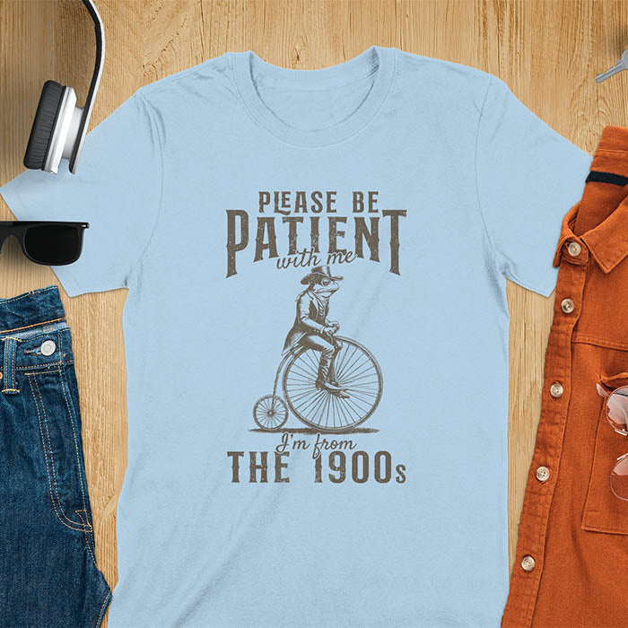 A light blue t-shirt with a frog riding a penny-farthing bicycle. Text: "PLEASE BE PATIENT with me" above and "I'm from THE 1900s" below. This The 1900s shirt has a vintage graphic tee and retro aesthetic. A genz Meme shirt and weird silly shirt, a funny gift friend. A gift for husband, funny grandpa gift, or dad tee-shirt. An animal person gift, a 90s country shirt, a mom gift option.