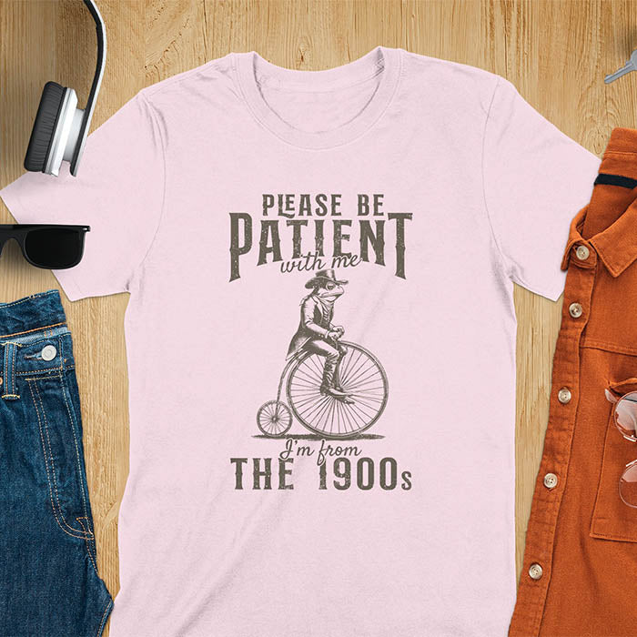 A light pink t-shirt with a frog riding a penny-farthing bicycle. Text: "PLEASE BE PATIENT with me" above and "I'm from THE 1900s" below. This The 1900s shirt has a vintage graphic tee and retro aesthetic. A genz Meme shirt and weird silly shirt, a funny gift friend. A gift for husband, funny grandpa gift, or dad tee-shirt. An animal person gift, a 90s country shirt, a mom gift option.