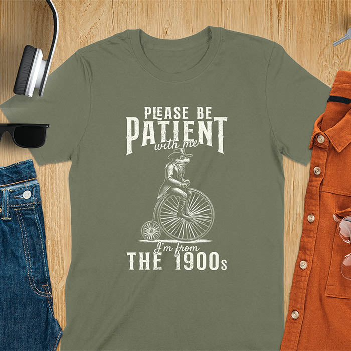 A military green t-shirt with a frog riding a penny-farthing bicycle. Text: "PLEASE BE PATIENT with me" above and "I'm from THE 1900s" below. This The 1900s shirt has a vintage graphic tee and retro aesthetic. A genz Meme shirt and weird silly shirt, a funny gift friend. A gift for husband, funny grandpa gift, or dad tee-shirt. An animal person gift, a 90s country shirt, a mom gift option.