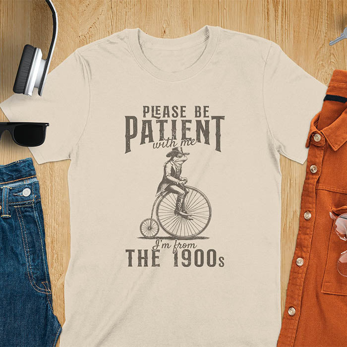 A sand t-shirt with a frog riding a penny-farthing bicycle. Text: "PLEASE BE PATIENT with me" above and "I'm from THE 1900s" below. This The 1900s shirt has a vintage graphic tee and retro aesthetic. A genz Meme shirt and weird silly shirt, a funny gift friend. A gift for husband, funny grandpa gift, or dad tee-shirt. An animal person gift, a 90s country shirt, a mom gift option.