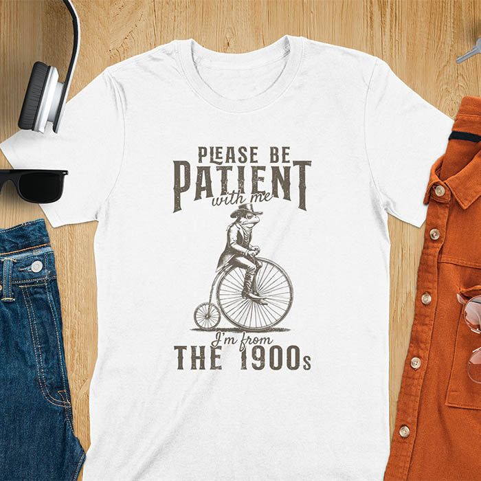 A white t-shirt with a frog riding a penny-farthing bicycle. Text: "PLEASE BE PATIENT with me" above and "I'm from THE 1900s" below. This The 1900s shirt has a vintage graphic tee and retro aesthetic. A genz Meme shirt and weird silly shirt, a funny gift friend. A gift for husband, funny grandpa gift, or dad tee-shirt. An animal person gift, a 90s country shirt, a mom gift option.