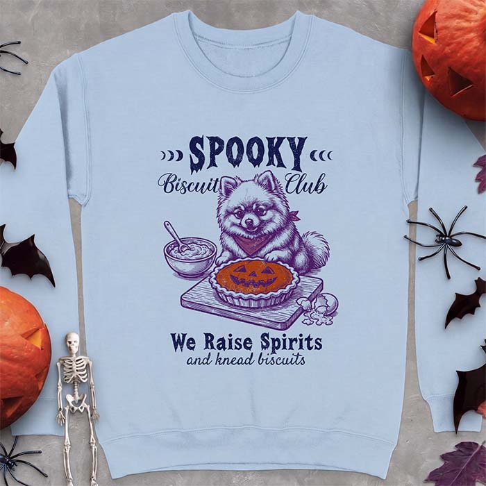 Light Blue sweatshirt with a Halloween graphic of a Pomeranian wearing a plaid bandana sitting behind a jack-o'-lantern pumpkin pie on a cutting board with a mixing bowl, spoon and a cracked egg nearby and the text Spooky Biscuit Club, We Raise Spirits and knead biscuits