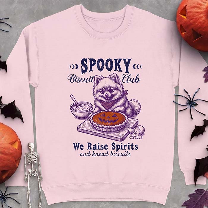 Light Pink sweatshirt with a Halloween graphic of a Pomeranian wearing a plaid bandana sitting behind a jack-o'-lantern pumpkin pie on a cutting board with a mixing bowl, spoon and a cracked egg nearby and the text Spooky Biscuit Club, We Raise Spirits and knead biscuits