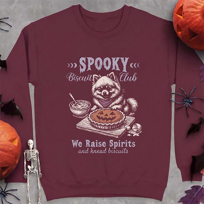 Maroon sweatshirt with a Halloween graphic of a Pomeranian wearing a plaid bandana sitting behind a jack-o'-lantern pumpkin pie on a cutting board with a mixing bowl, spoon and a cracked egg nearby and the text Spooky Biscuit Club, We Raise Spirits and knead biscuits