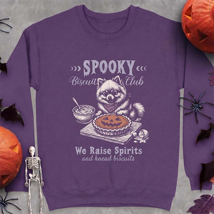 Purple sweatshirt with a Halloween graphic of a Pomeranian wearing a plaid bandana sitting behind a jack-o'-lantern pumpkin pie on a cutting board with a mixing bowl, spoon and a cracked egg nearby and the text Spooky Biscuit Club, We Raise Spirits and knead biscuits