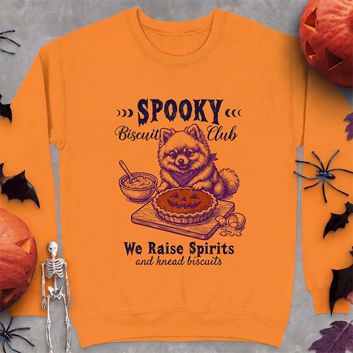 Safety Orange sweatshirt with a Halloween graphic of a Pomeranian wearing a plaid bandana sitting behind a jack-o'-lantern pumpkin pie on a cutting board with a mixing bowl, spoon and a cracked egg nearby and the text Spooky Biscuit Club, We Raise Spirits and knead biscuits