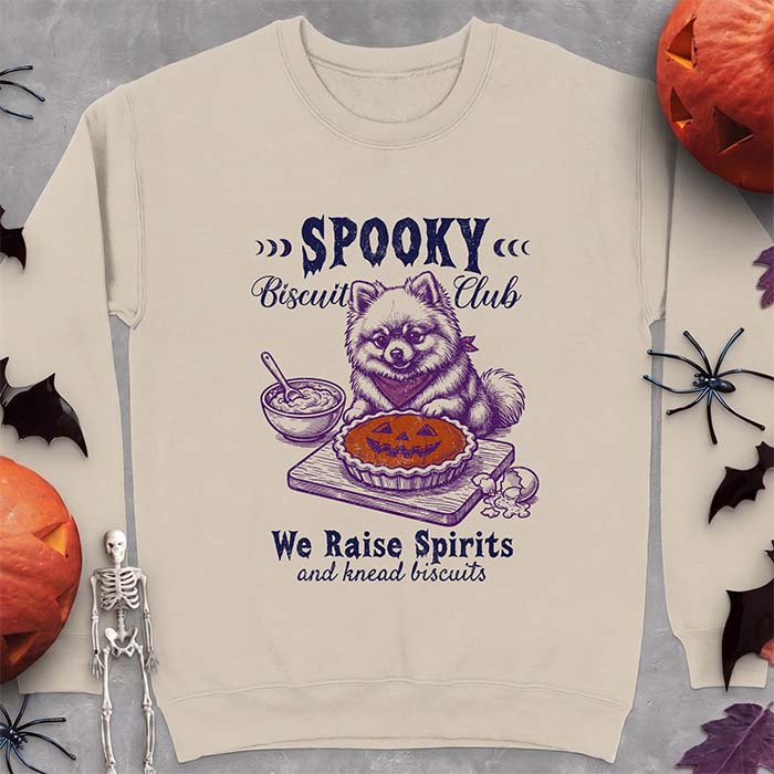 Sand sweatshirt with a Halloween graphic of a Pomeranian wearing a plaid bandana sitting behind a jack-o'-lantern pumpkin pie on a cutting board with a mixing bowl, spoon and a cracked egg nearby and the text Spooky Biscuit Club, We Raise Spirits and knead biscuits