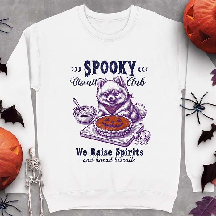 White sweatshirt with a Halloween graphic of a Pomeranian wearing a plaid bandana sitting behind a jack-o'-lantern pumpkin pie on a cutting board with a mixing bowl, spoon and a cracked egg nearby and the text Spooky Biscuit Club, We Raise Spirits and knead biscuits