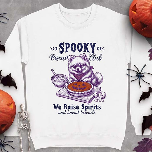 White sweatshirt with a Halloween graphic of a Pomeranian wearing a plaid bandana sitting behind a jack-o'-lantern pumpkin pie on a cutting board with a mixing bowl, spoon and a cracked egg nearby and the text Spooky Biscuit Club, We Raise Spirits and knead biscuits