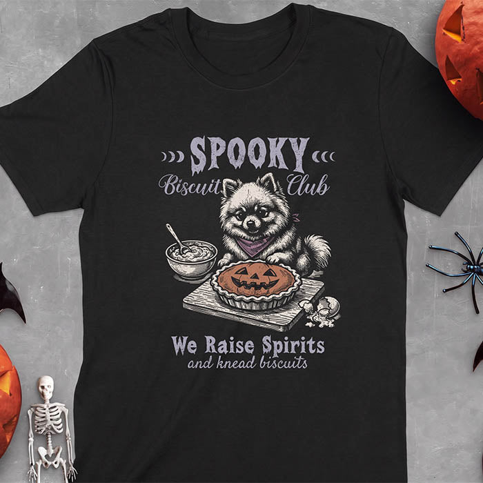 Black t-shirt with a Halloween graphic of a Pomeranian wearing a plaid bandana sitting behind a jack-o'-lantern pumpkin pie on a cutting board with a mixing bowl, spoon and a cracked egg nearby and the text Spooky Biscuit Club, We Raise Spirits and knead biscuits