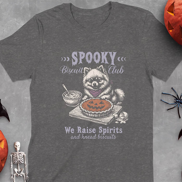 Dark Heather t-shirt with a Halloween graphic of a Pomeranian wearing a plaid bandana sitting behind a jack-o'-lantern pumpkin pie on a cutting board with a mixing bowl, spoon and a cracked egg nearby and the text Spooky Biscuit Club, We Raise Spirits and knead biscuits
