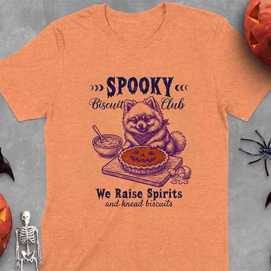 Heather Orange t-shirt with a Halloween graphic of a Pomeranian wearing a plaid bandana sitting behind a jack-o'-lantern pumpkin pie on a cutting board with a mixing bowl, spoon and a cracked egg nearby and the text Spooky Biscuit Club, We Raise Spirits and knead biscuits