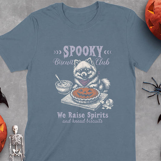 Indigo Blue t-shirt with a Halloween graphic of a Pomeranian wearing a plaid bandana sitting behind a jack-o'-lantern pumpkin pie on a cutting board with a mixing bowl, spoon and a cracked egg nearby and the text Spooky Biscuit Club, We Raise Spirits and knead biscuits