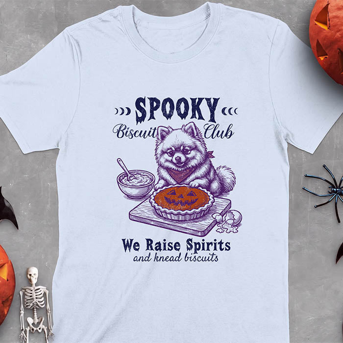 Light Blue t-shirt with a Halloween graphic of a Pomeranian wearing a plaid bandana sitting behind a jack-o'-lantern pumpkin pie on a cutting board with a mixing bowl, spoon and a cracked egg nearby and the text Spooky Biscuit Club, We Raise Spirits and knead biscuits