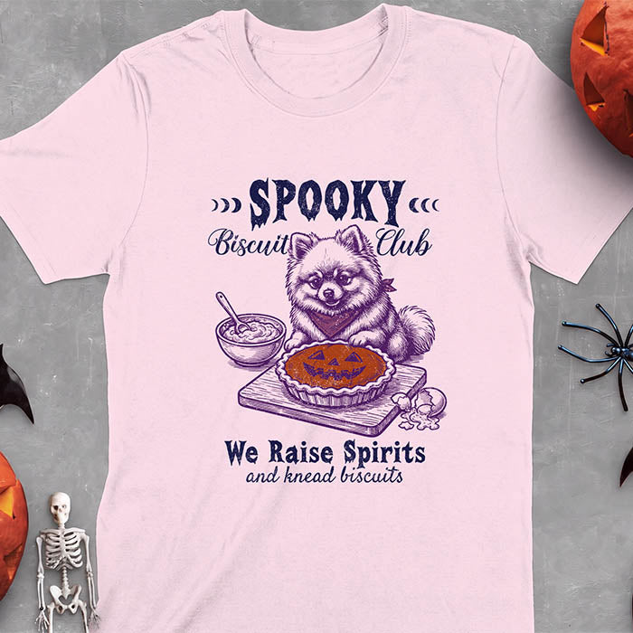 Light Pink t-shirt with a Halloween graphic of a Pomeranian wearing a plaid bandana sitting behind a jack-o'-lantern pumpkin pie on a cutting board with a mixing bowl, spoon and a cracked egg nearby and the text Spooky Biscuit Club, We Raise Spirits and knead biscuits