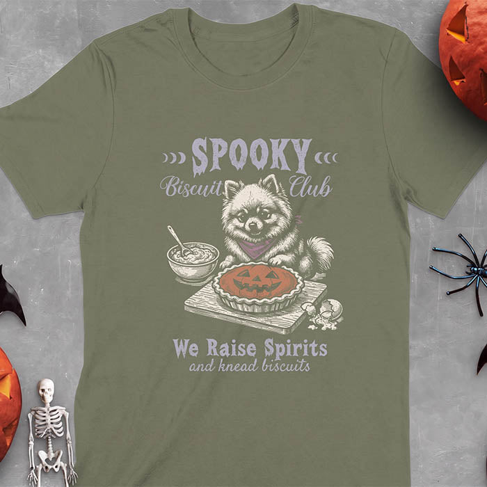 Military Green t-shirt with a Halloween graphic of a Pomeranian wearing a plaid bandana sitting behind a jack-o'-lantern pumpkin pie on a cutting board with a mixing bowl, spoon and a cracked egg nearby and the text Spooky Biscuit Club, We Raise Spirits and knead biscuits