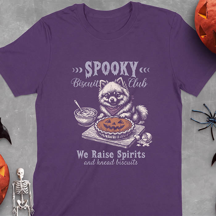 Purple t-shirt with a Halloween graphic of a Pomeranian wearing a plaid bandana sitting behind a jack-o'-lantern pumpkin pie on a cutting board with a mixing bowl, spoon and a cracked egg nearby and the text Spooky Biscuit Club, We Raise Spirits and knead biscuits
